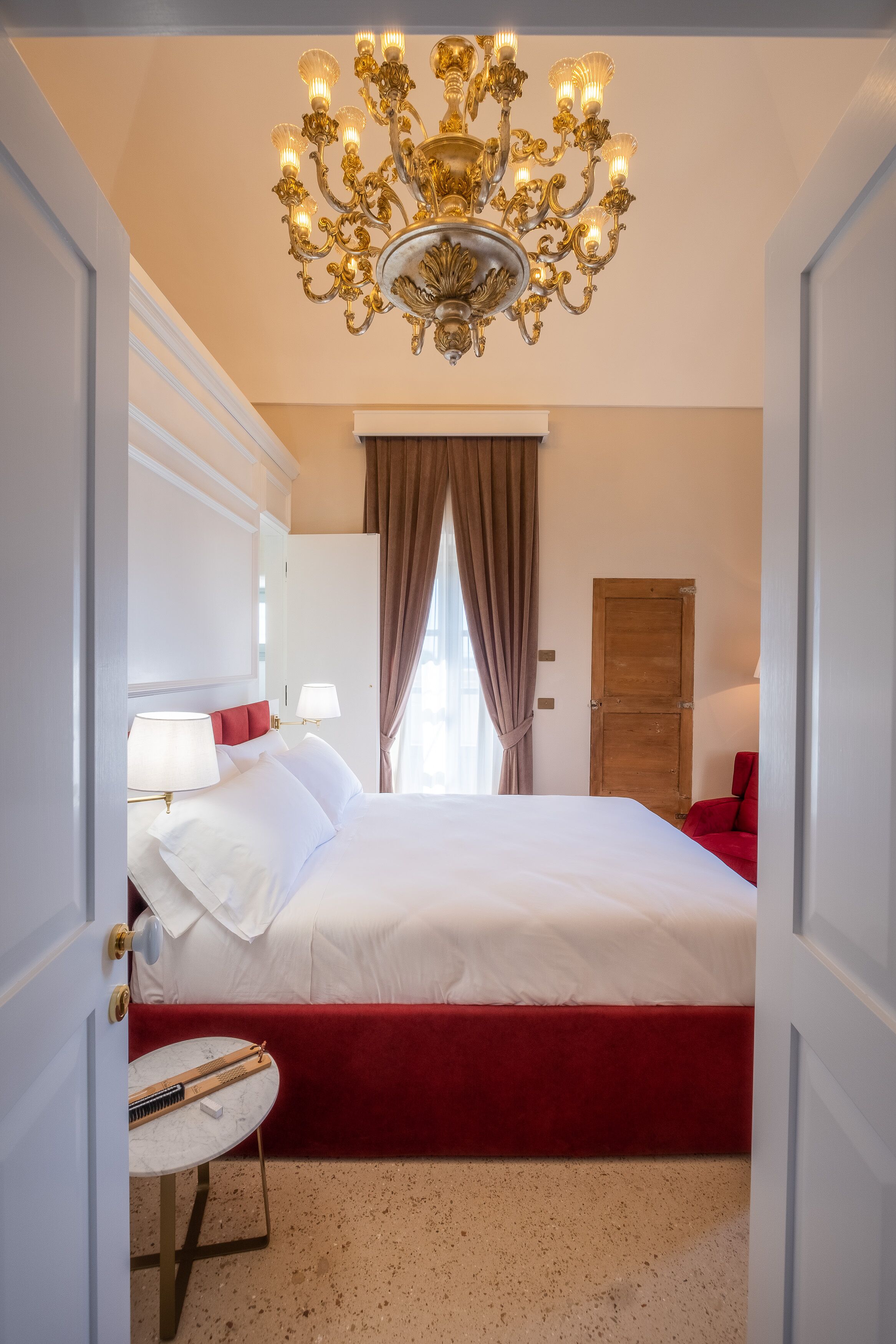 A hotel room with a large bed, a chandelier, a red upholstered bench, and a small round table with a lamp.
