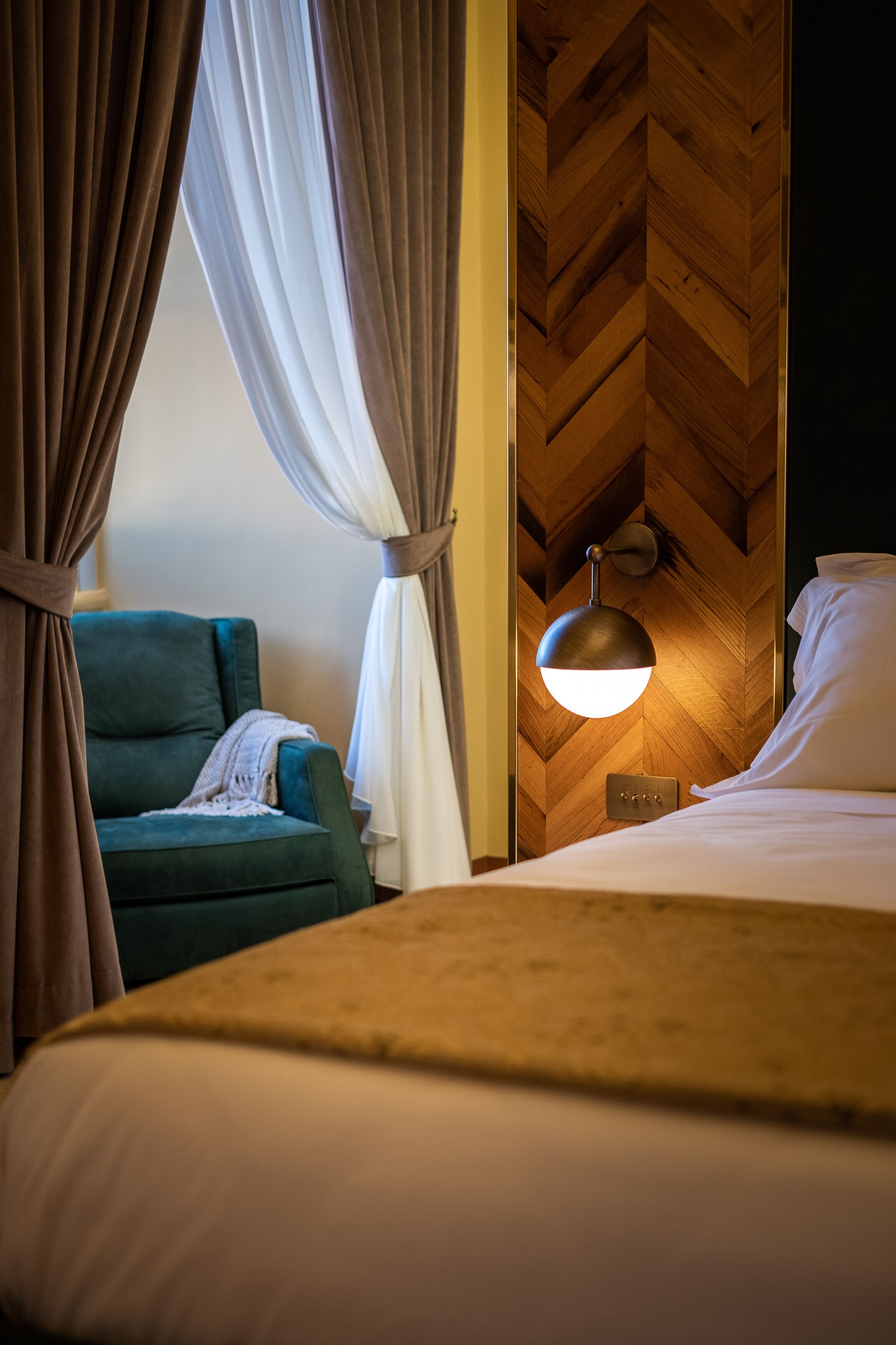 Ein Hotelzimmer mit einem Bett, einem grünen Sessel, einer Wandleuchte und Vorhängen.
