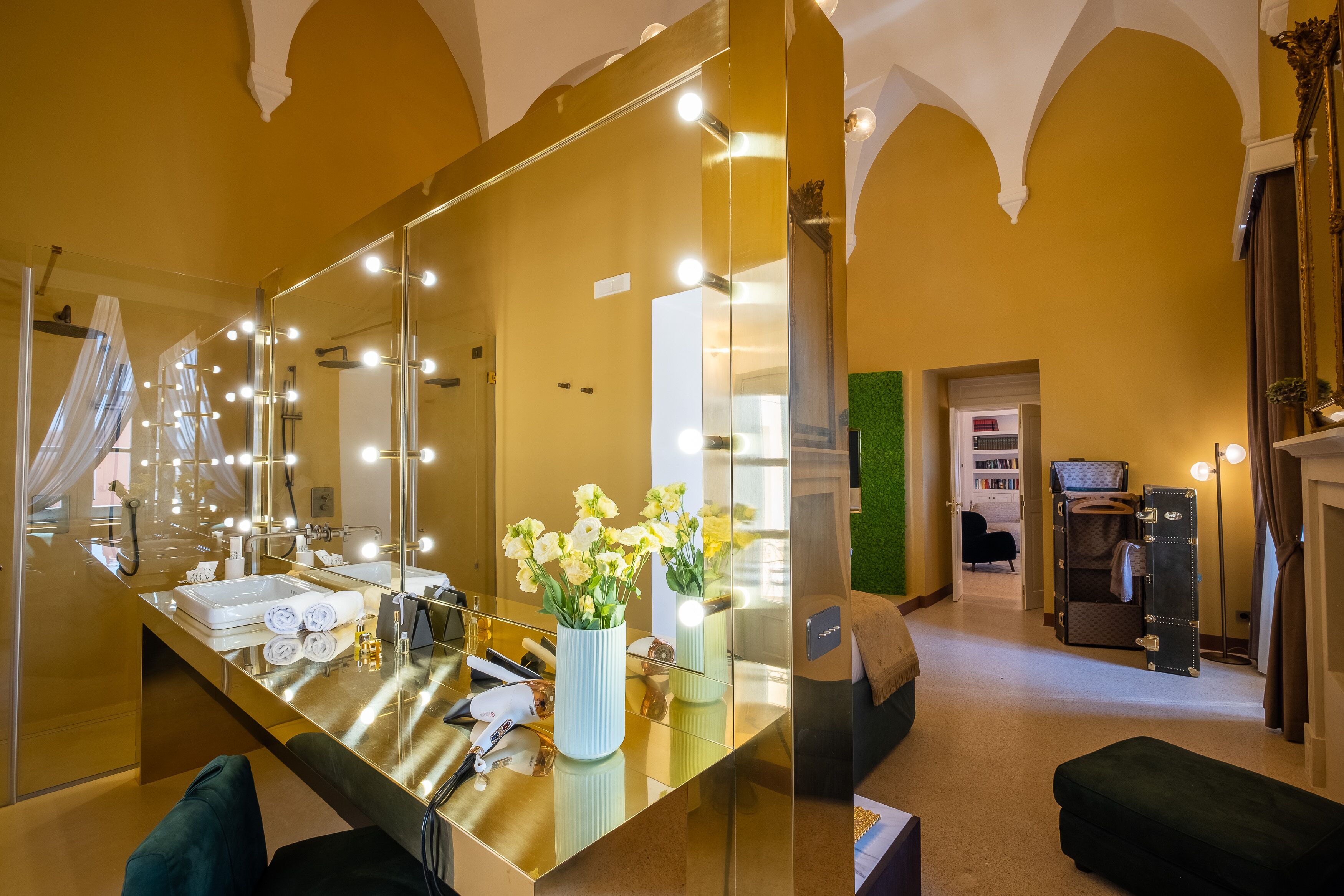A hotel room with a large mirror, vanity with lights, a bed, and a green wall.
