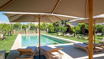 Un espace aménagé au bord de la piscine, avec des chaises longues et des parasols, entouré de verdure et d’un mur en pierre.