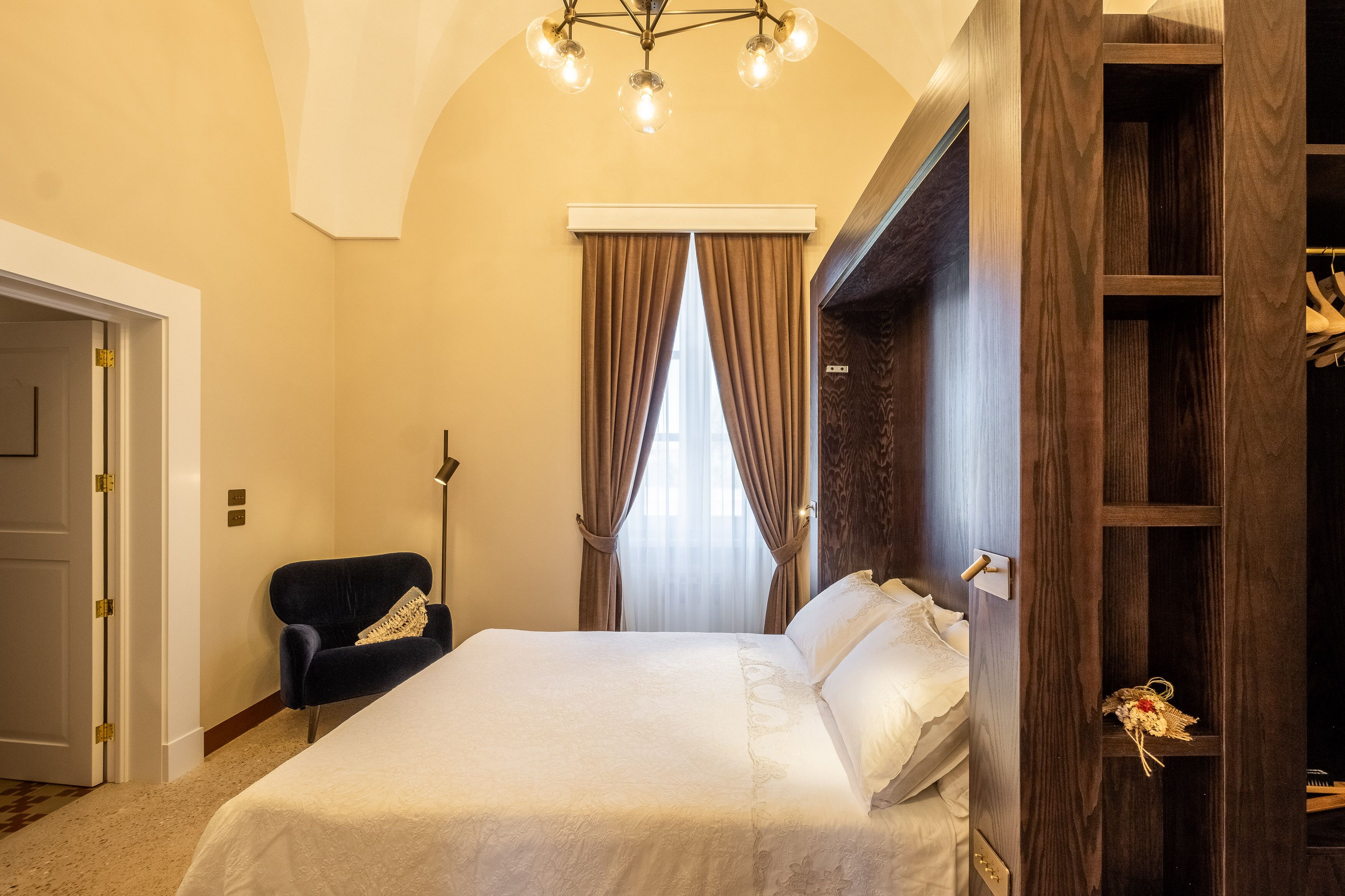 A hotel room with a large bed, a dark armchair, a wooden wardrobe, and a chandelier.