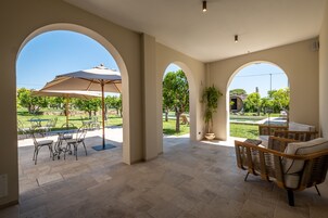 A covered patio area with outdoor seating, a potted plant, and a view of a garden and pool.