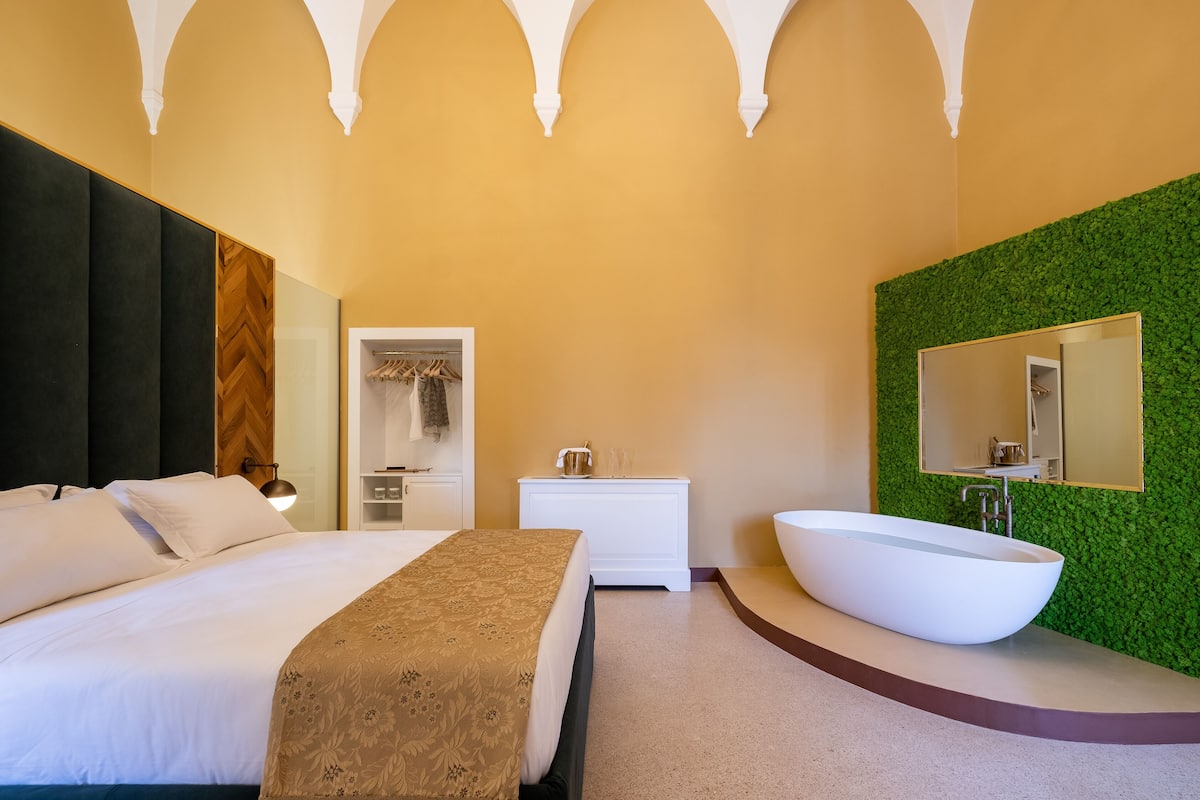Una habitación de hotel moderna con una cama grande, una mesita de noche blanca, un banco empotrado, una bañera blanca amplia y una pared verde con espejo.