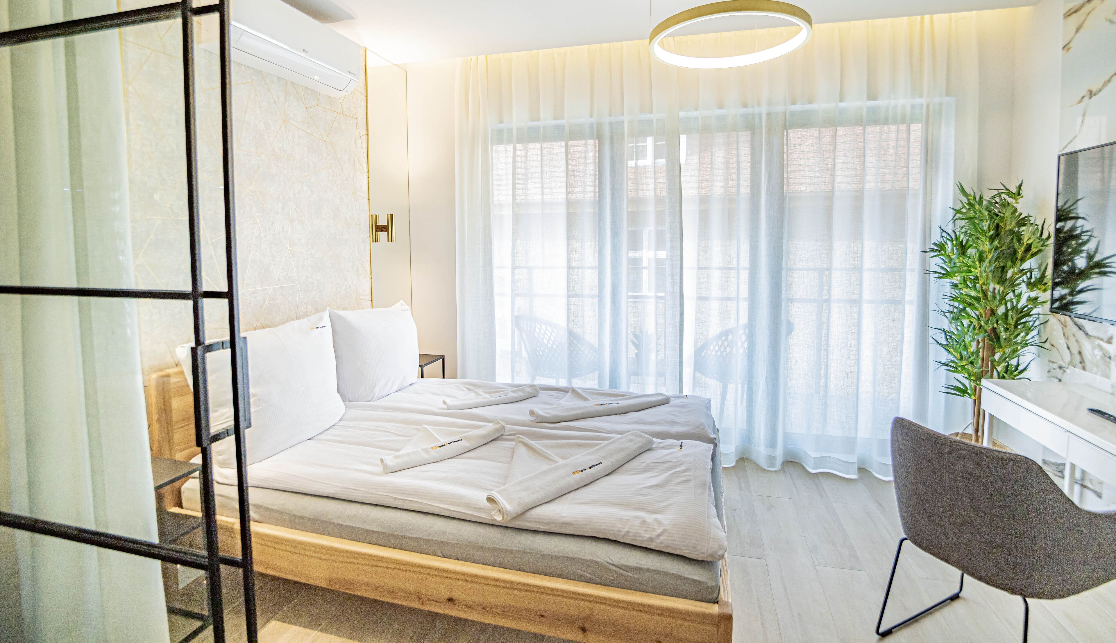 Egy modern hálószoba, melyben egy faágy, fehér ágynemű, egy szürke szék, egy növény és egy fehér szekrényen elhelyezett televízió található.