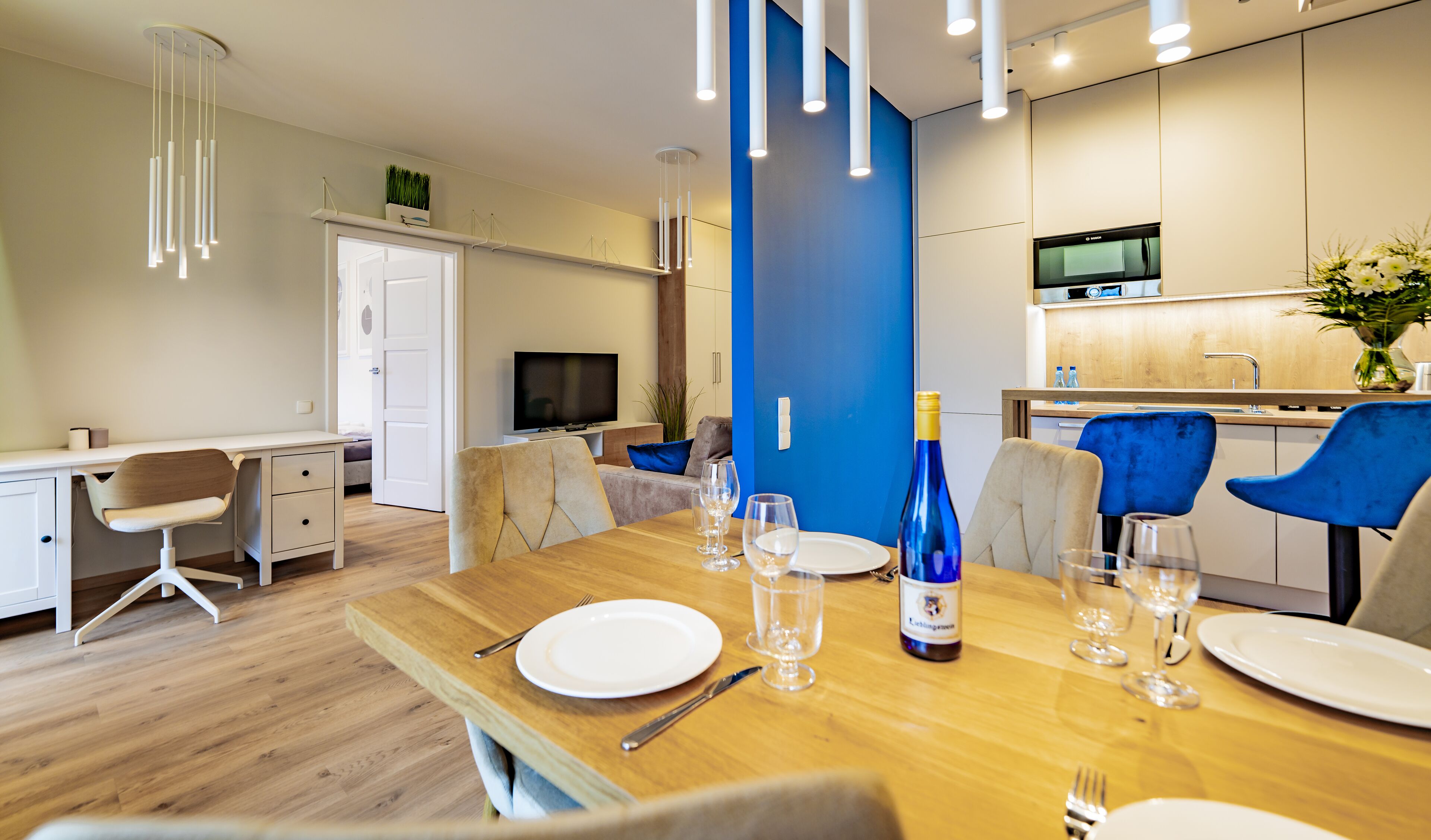 Un espace de repas moderne avec une table en bois dressée pour le repas, un téléviseur en arrière-plan et un mur d’accent bleu.