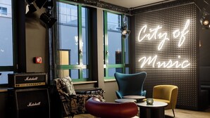Una habitación con un letrero de neón que dice "City of Music", un amplificador Marshall, un sillón estampado y una pequeña mesa redonda.