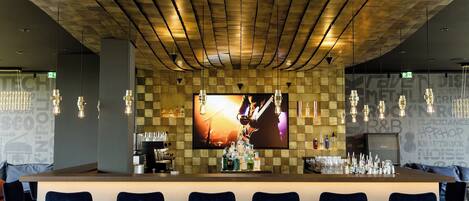 Un espace bar avec un plafond à carreaux, un grand écran diffusant des images d’un musicien et des tabourets de bar.