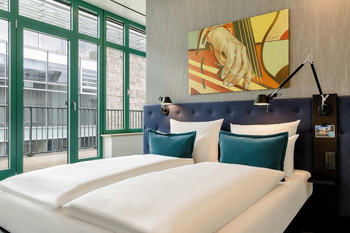 Una habitación de hotel con una cama, cabecera, un cuadro y una mesita de noche con lámpara.