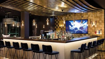 Zona de bar con una barra larga, varios taburetes y una pantalla de televisión mostrando una banda.