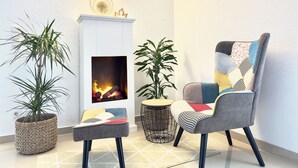 Een gezellige woonkamer met een open haard, twee potplanten, een gestreept fauteuil en een klein rond tafeltje.