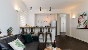 Ein modernes Wohnzimmer mit einer Ledersofa, einem Holztisch mit einer Flasche und Gläsern, sowie einer Küche im Hintergrund mit Barhocker und einem Kühlschrank.