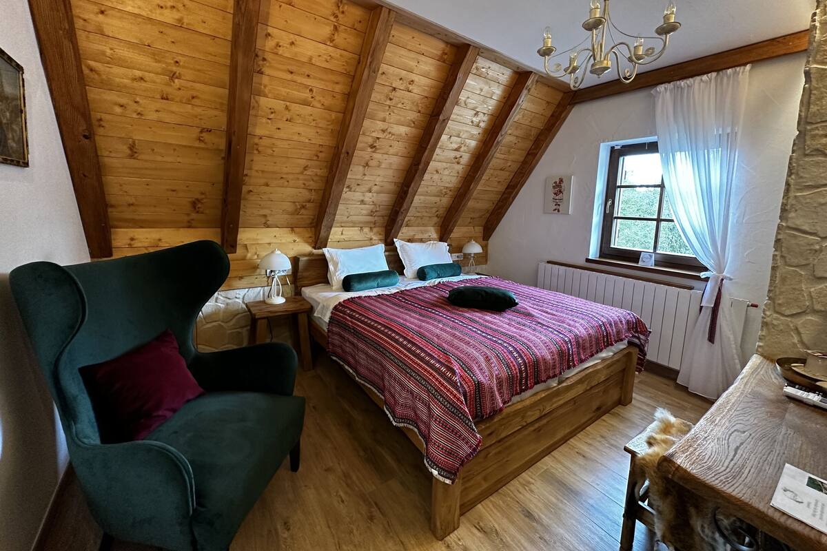 Ein gemütliches Schlafzimmer im Dachgeschoss mit Holzbalkendecke, einem Bett mit gemustertem Überwurf, einem Sessel und einem Tisch mit Lampe.