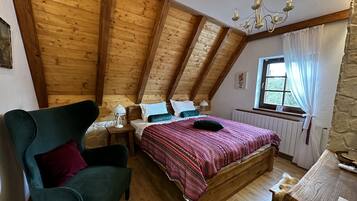 A cozy attic bedroom with a wooden ceiling, a bed with a patterned blanket, a chair, and a table with a lamp.
