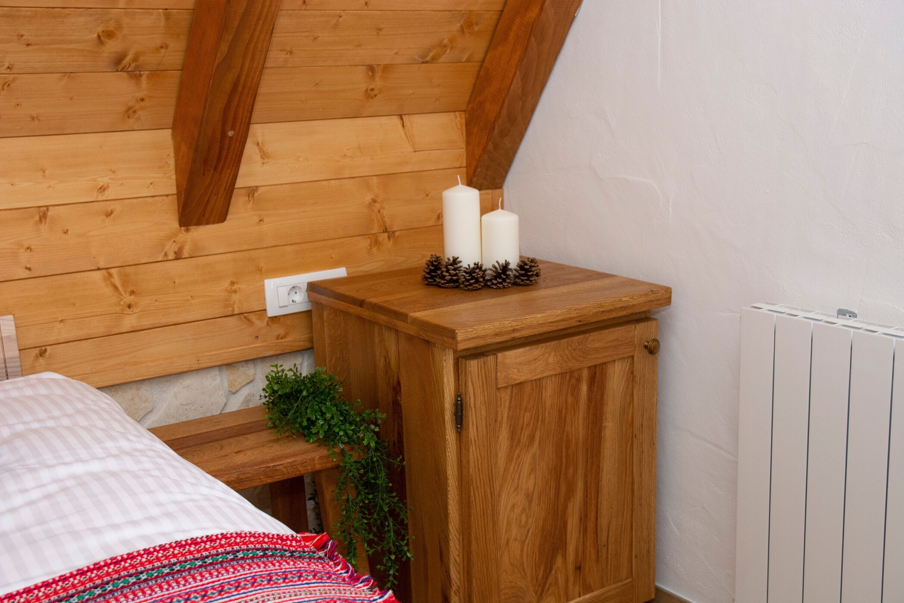 Un mueble de madera con dos velas blancas y piñas encima, una cama con manta estampada y un radiador en la esquina.