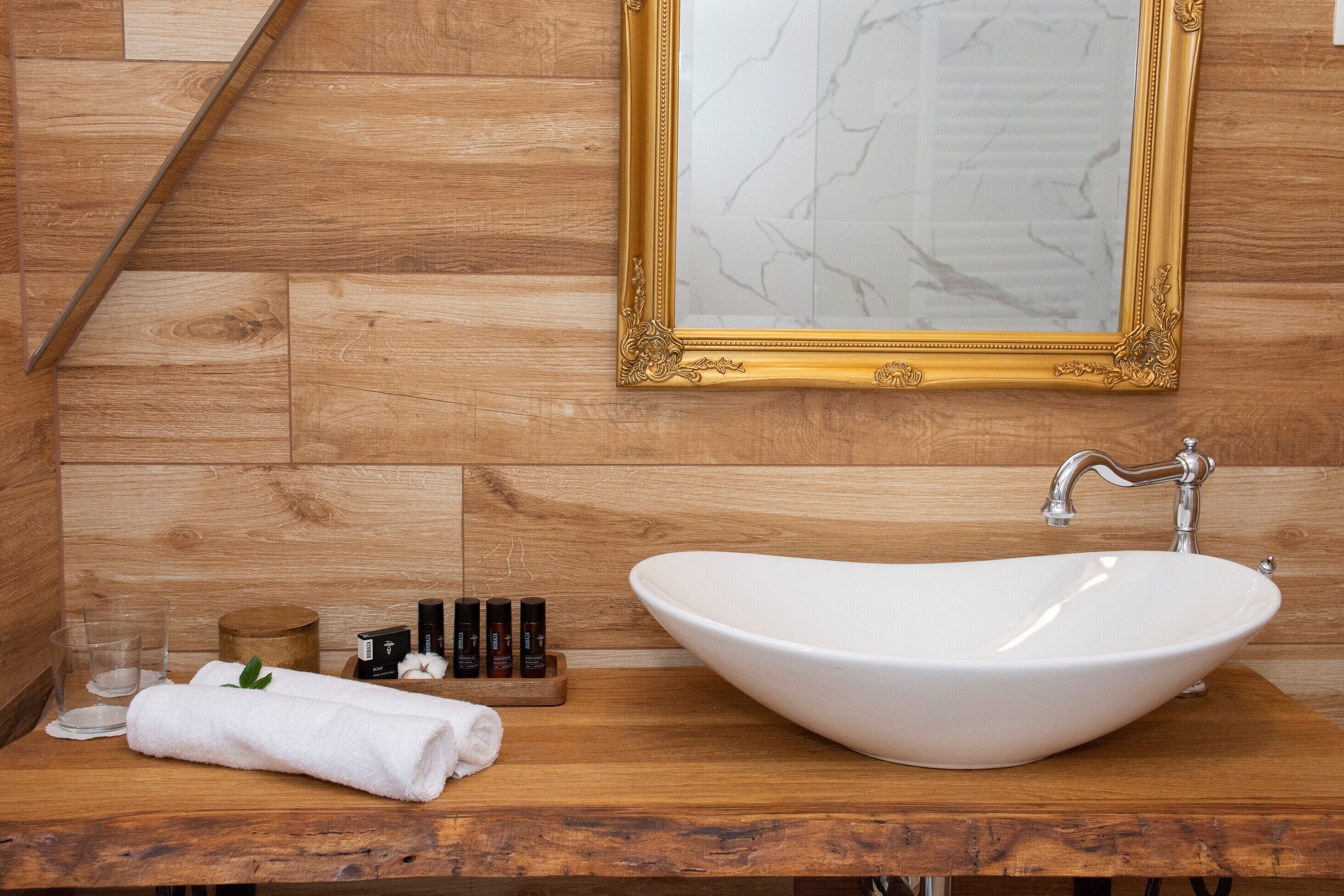 Une salle de bain avec un meuble-lavabo en bois, un lavabo blanc, un miroir à cadre doré et un mur en bois.