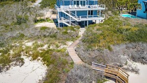 Una casa grande de dos pisos frente a la playa, con un porche cubierto y un camino de madera que conduce a la entrada.