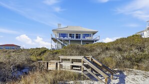 Una casa frente a la playa con balcón, rodeada de vegetación costera y una escalera de madera que lleva a la entrada.