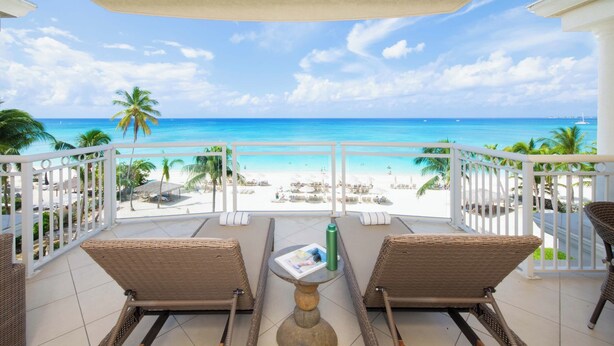Een balkon met rieten stoelen en een tafel, met uitzicht op een strand met palmbomen en een helderblauwe lucht.