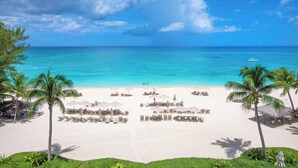 Een strand met helder turquoise water, wit zand en palmbomen. Er staan ligbedden en parasols op het strand.