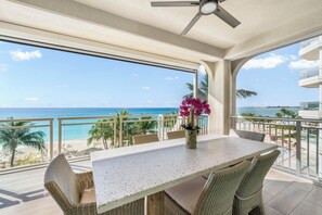 En balkon med bord og stole, der vender ud mod en strand og et hav.