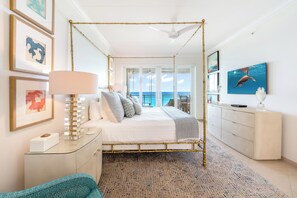 Un lit à baldaquin avec vue sur l’océan, une commode blanche, une lampe et des œuvres d’art encadrées accrochées au mur.