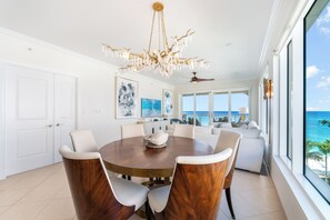 Un vaste espace dédié à la salle à manger, avec une table ronde en bois et des chaises à revêtement blanc, un lustre et une vue sur l’océan grâce à de grandes fenêtres.