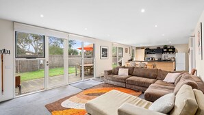 A spacious living room with a large sliding glass door leading to a backyard, a cozy brown sofa, and a kitchen area with modern appliances. - Spindrift Point Lonsdale (Point Lonsdale)