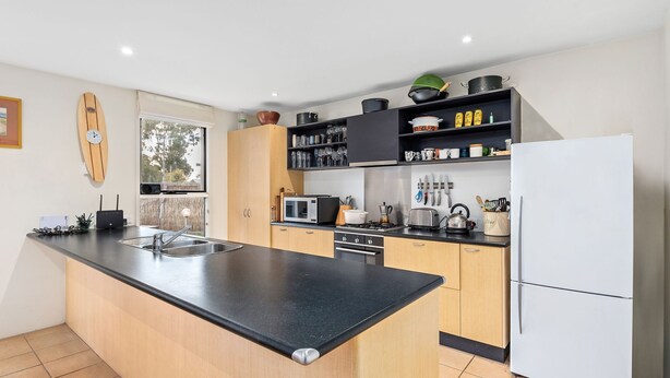 A modern kitchen with a central island, black countertops, wooden cabinets, and a white refrigerator. - Spindrift Point Lonsdale (Point Lonsdale)