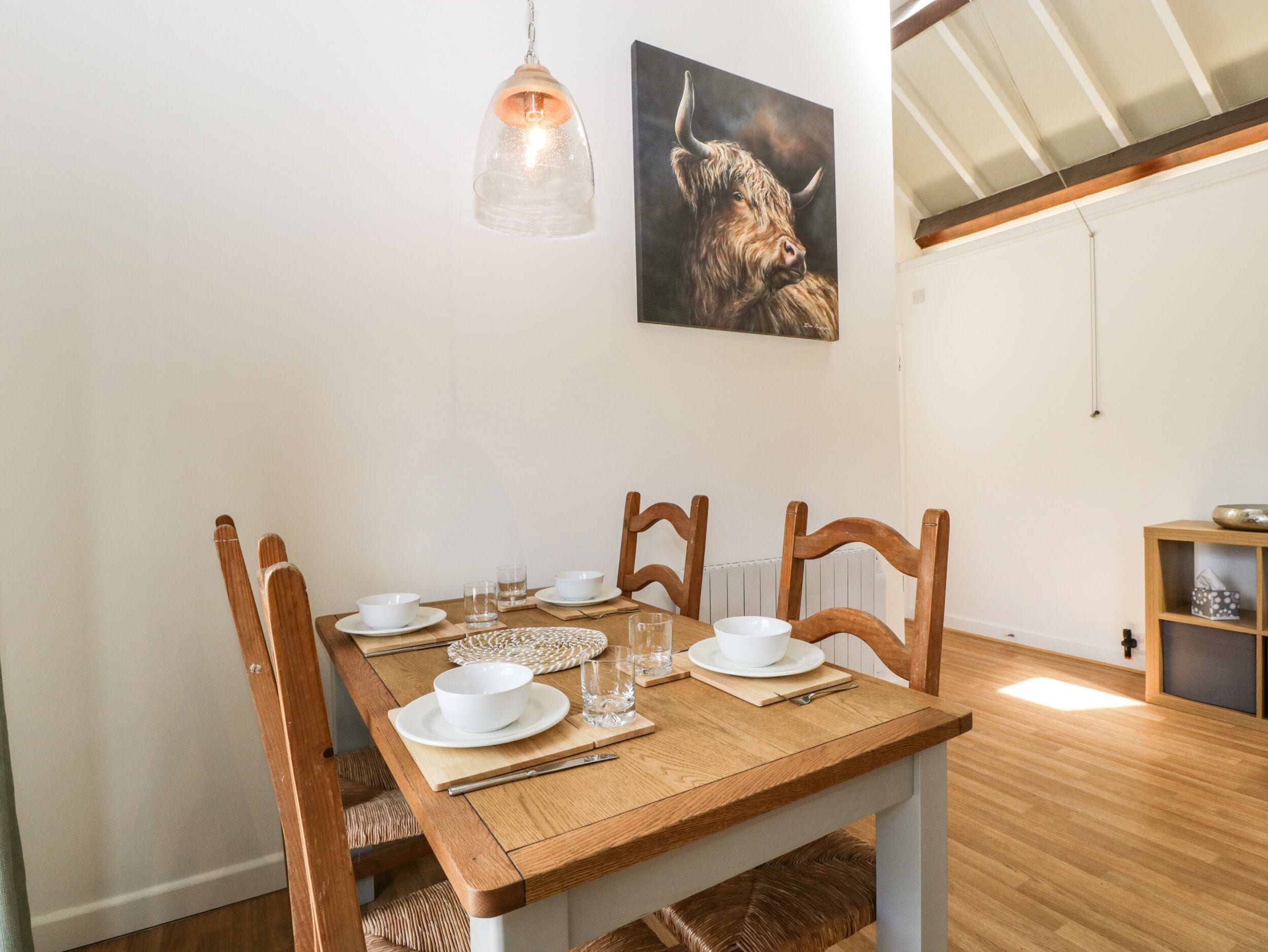 A dining area with a wooden table set for four, surrounded by wooden chairs. A pendant light hangs above the table, and a painting of a bull is on the wall.