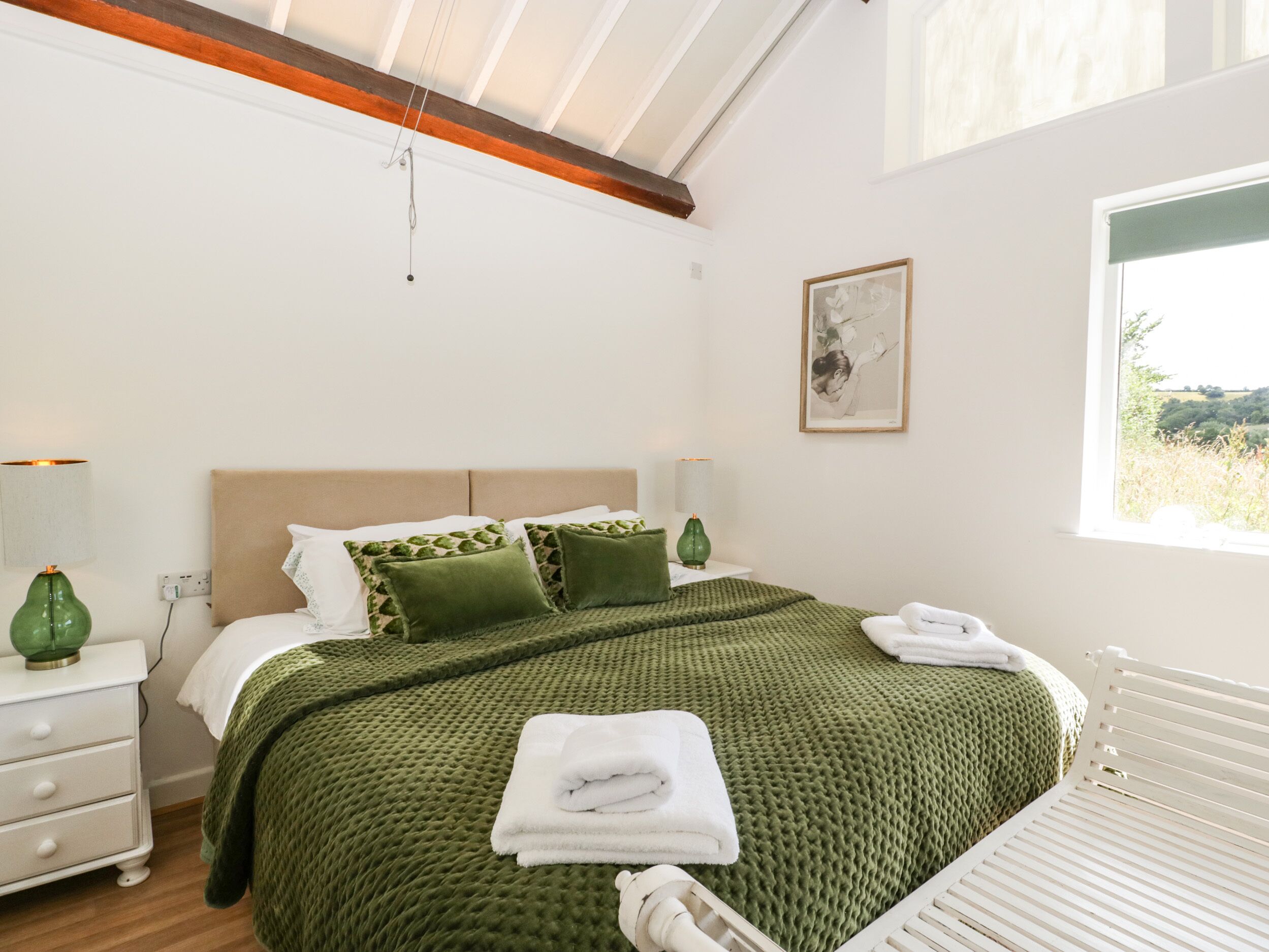A bedroom with a large bed, green bedding, a white dresser, a bedside table with a lamp, and a window with a view of greenery.
