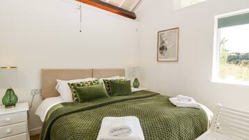 A bedroom with a large bed, green bedding, a white dresser, a bedside table with a lamp, and a window with a view of greenery.