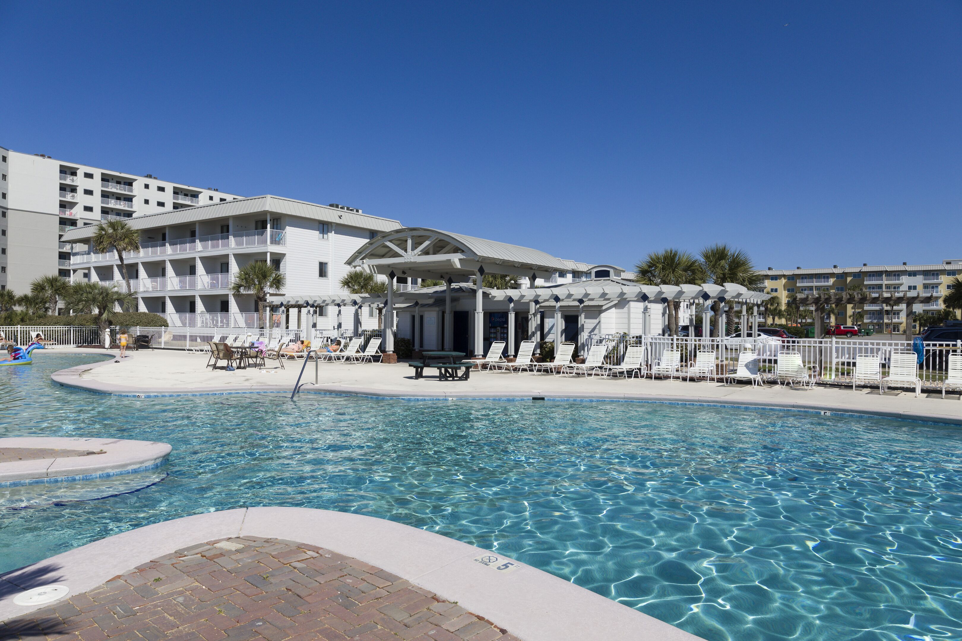 A hotel pool area with multiple lounge chairs and umbrellas, a covered seating area, and a clear blue pool.
