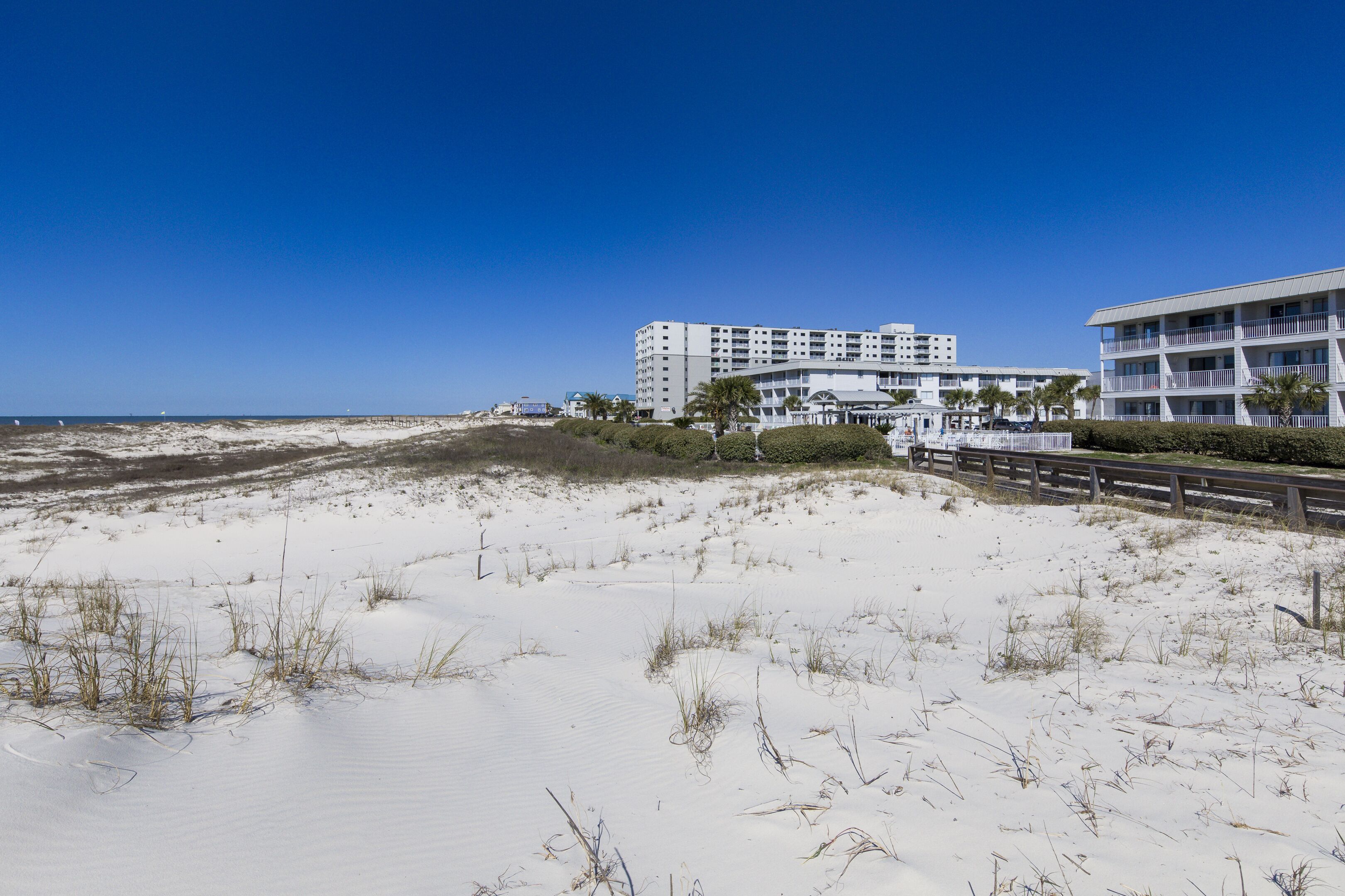 Un hotel frente a la playa con varios pisos, una playa arenosa en primer plano y un cielo azul despejado.