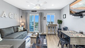 A modern living room with a grey sofa, a wooden coffee table, and a dining area with a wooden table and chairs. A ceiling fan is visible, and there is a TV mounted on the wall. - Plantation 4514 (Gulf Shores)