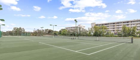Una cancha de tenis al aire libre con una red, rodeada de árboles y un edificio al fondo.