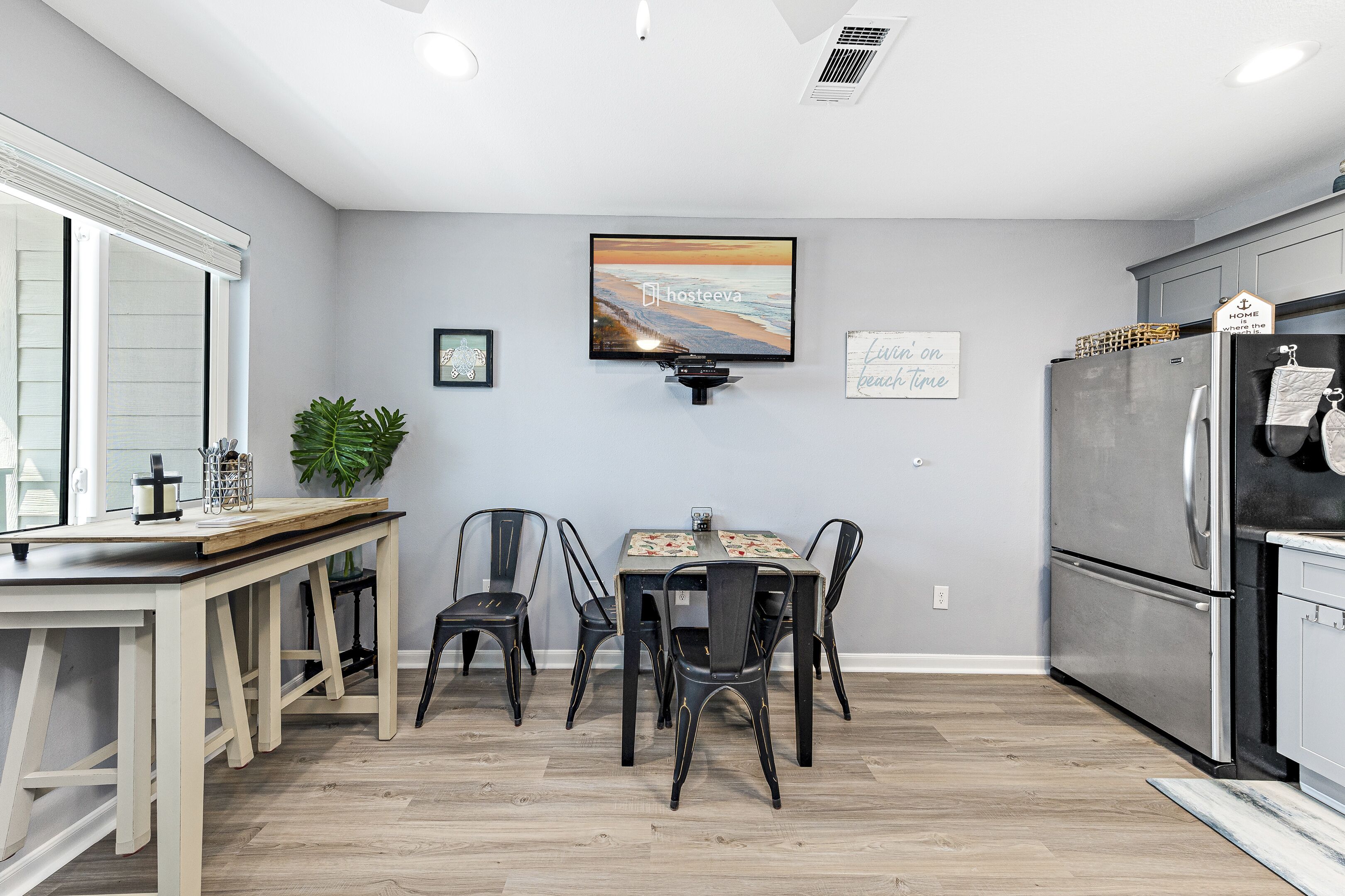 Een moderne keuken met een eethoek, met een grote flatscreen televisie, een koelkast en een houten eettafel met stoelen.
