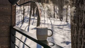 En hand häller vatten från en svart vattenkanna ner i en keramisk mugg som står på ett balkongräcke.