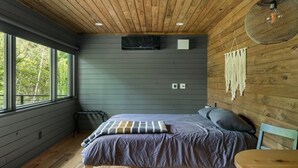 Un dormitorio con cama, una mesa redonda y vistas a árboles a través de amplios ventanales.