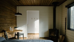 A cozy cabin room with a bed, a table with chairs, a wooden ceiling, a white door, and a wood-burning stove.