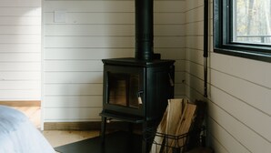 A room with a wood-burning stove, wooden flooring, and a window with a view of trees.