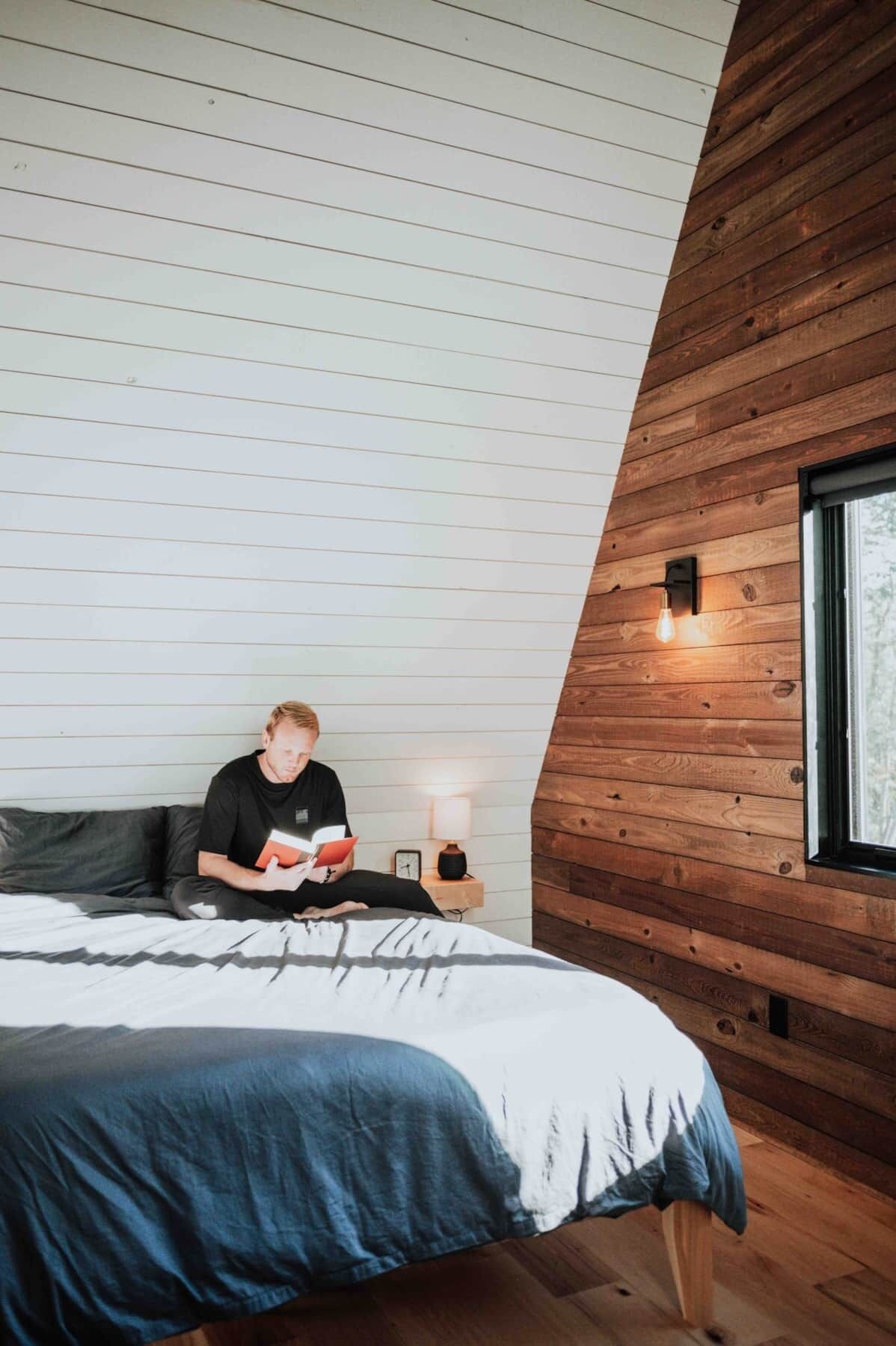 Ein Schlafzimmer mit schrägverdeckter Decke, einem Bett mit blauen und weißen Bettbezügen, einem Nachttisch mit Lampe und einem Fenster mit Blick auf Bäume.