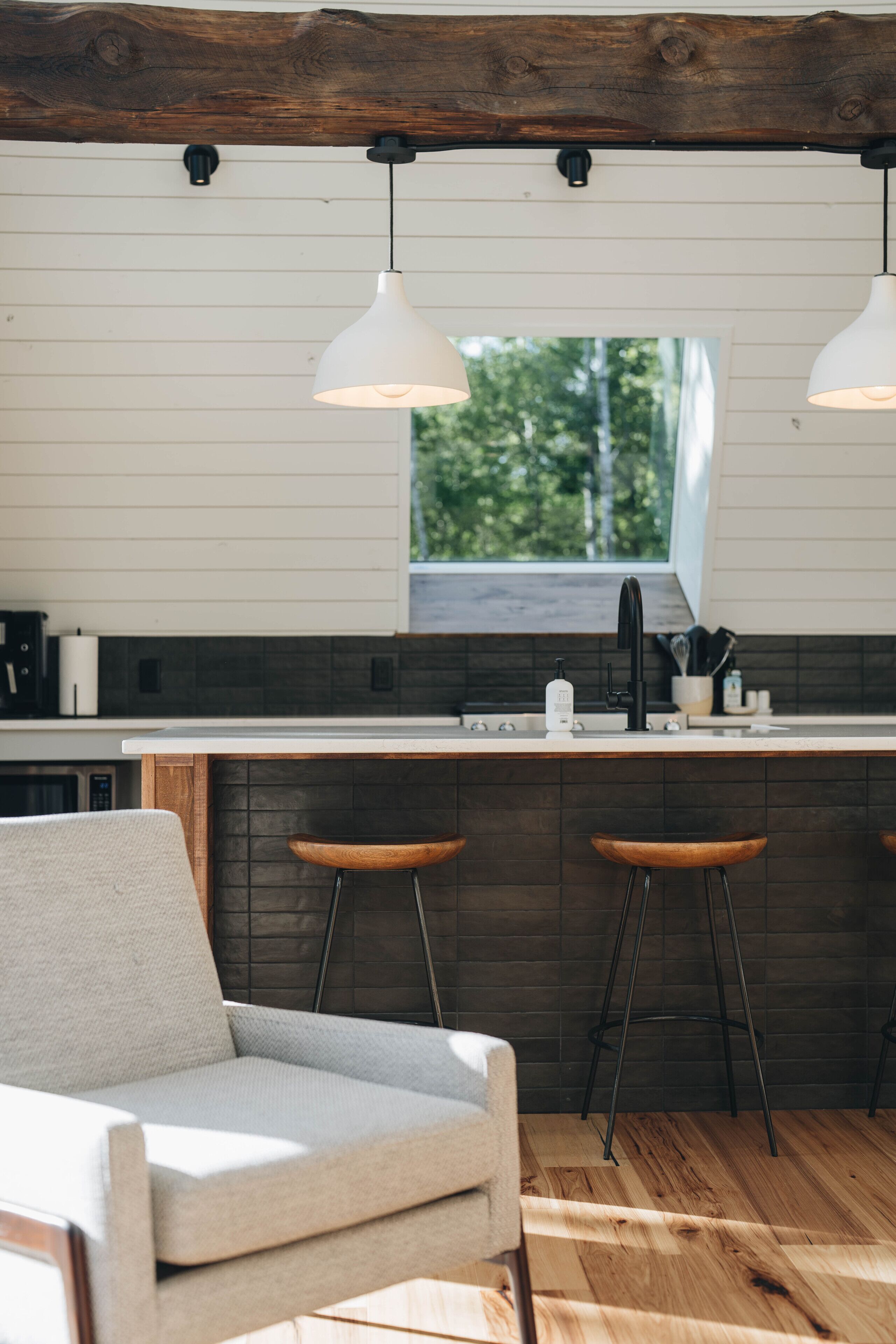 Una cocina moderna con techo de madera, paredes blancas y un ventanal. Hay dos taburetes de bar y una silla blanca.