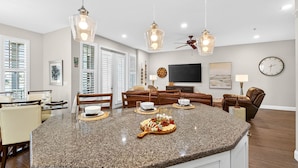 A spacious kitchen with granite countertops, a dining area with chairs, a living room with a sofa and a TV, and a ceiling fan.