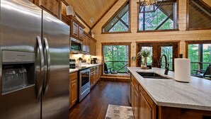 A modern kitchen with wooden cabinets, stainless steel appliances, and large windows overlooking a forested area. - HIGH Luxe MODERN 1+ Bedroom New Construction July 2022! (Broken Bow)