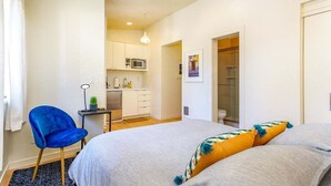 A bedroom with a bed, a blue chair, a small table with a lamp, and a kitchenette in the background.