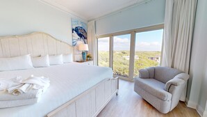 A bedroom with a large bed, a nightstand with a lamp, a chair, and a large window with a view of greenery and buildings.