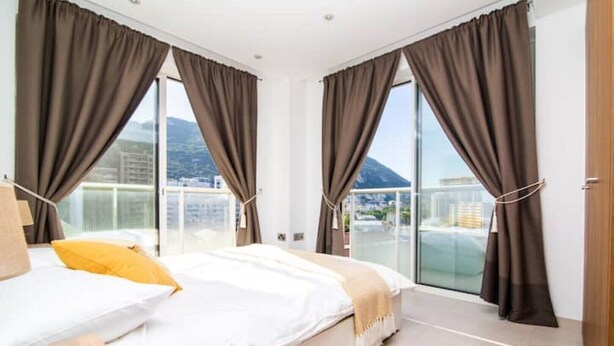 A modern hotel room with a large bed, two windows with curtains, a view of the city, and a balcony. - Luxury modern apartment with exceptional views! (Gibraltar)