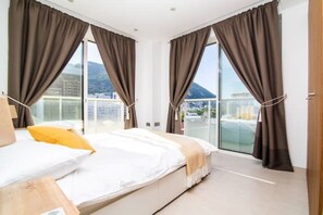 A modern hotel room with a large bed, two windows with curtains, a view of the city, and a balcony.