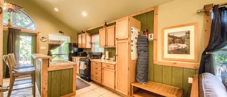 Cocina compacta con armarios de madera, estufa y lavabo. Hay un banco y una pequeña mesa.
