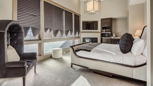 A modern bedroom with a large bed, a seating area with a chair, a small ottoman, and a built-in TV.